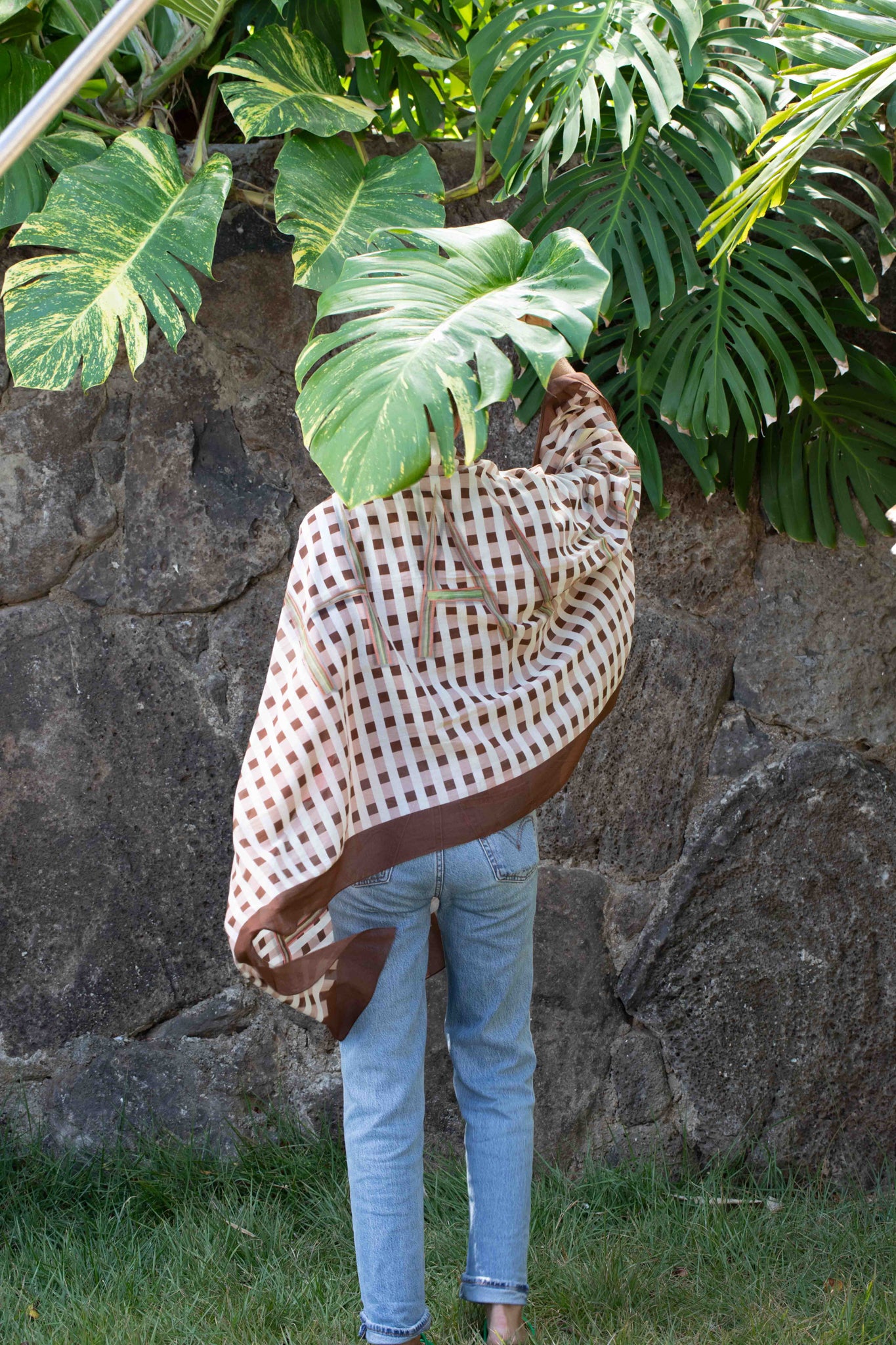 Person wearing a checkered shirt and jeans with a large leaf in front of a stone wall palaka style honolulu hawaii Lola Pilar